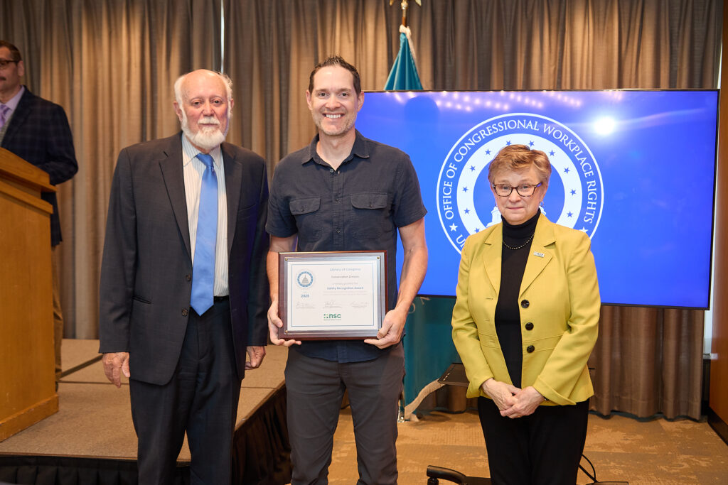 Three individuals stand side by side, the man in the middle holds a framed award