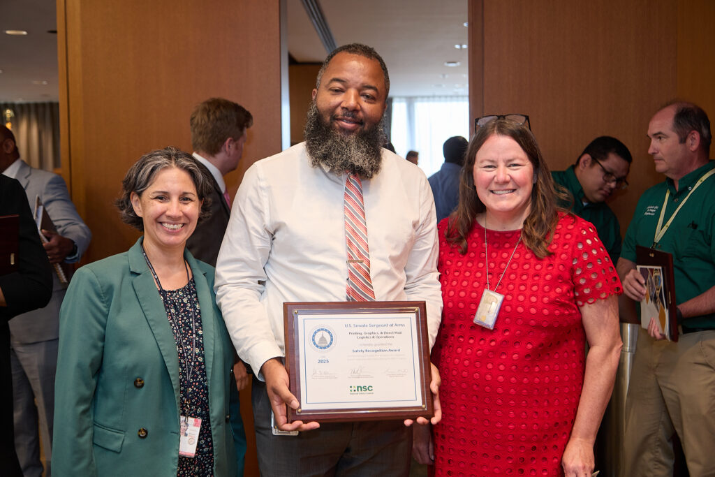 Group of three individuals, the man in the middle holds a framed award