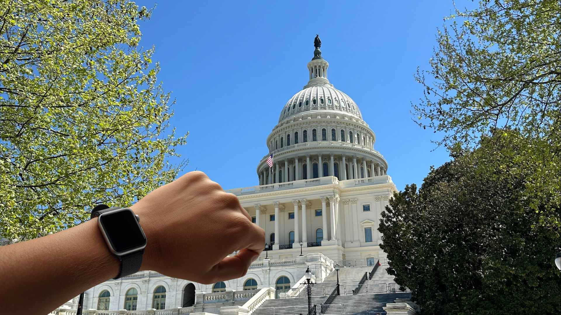 An arm with a smartwatch on its wrist is raised in front of the U.S. Capitol building