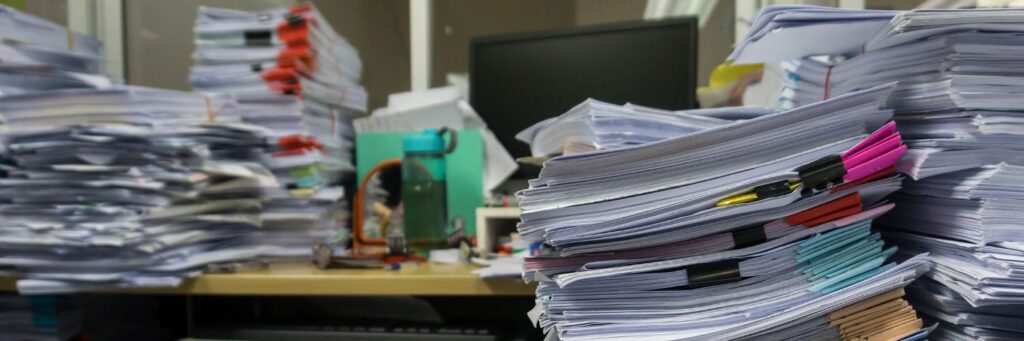 Haphazard stacks of paper surround a desk with a computer monitor and water bottle
