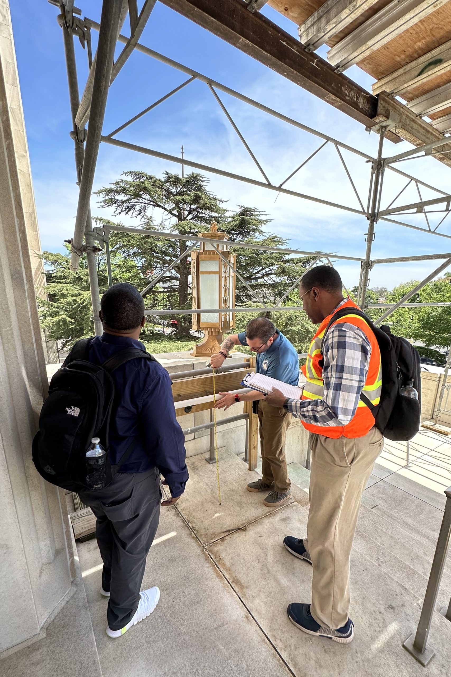 Three individuals measure the height of a railing on a staircase outside of a building that is covered in scaffolding