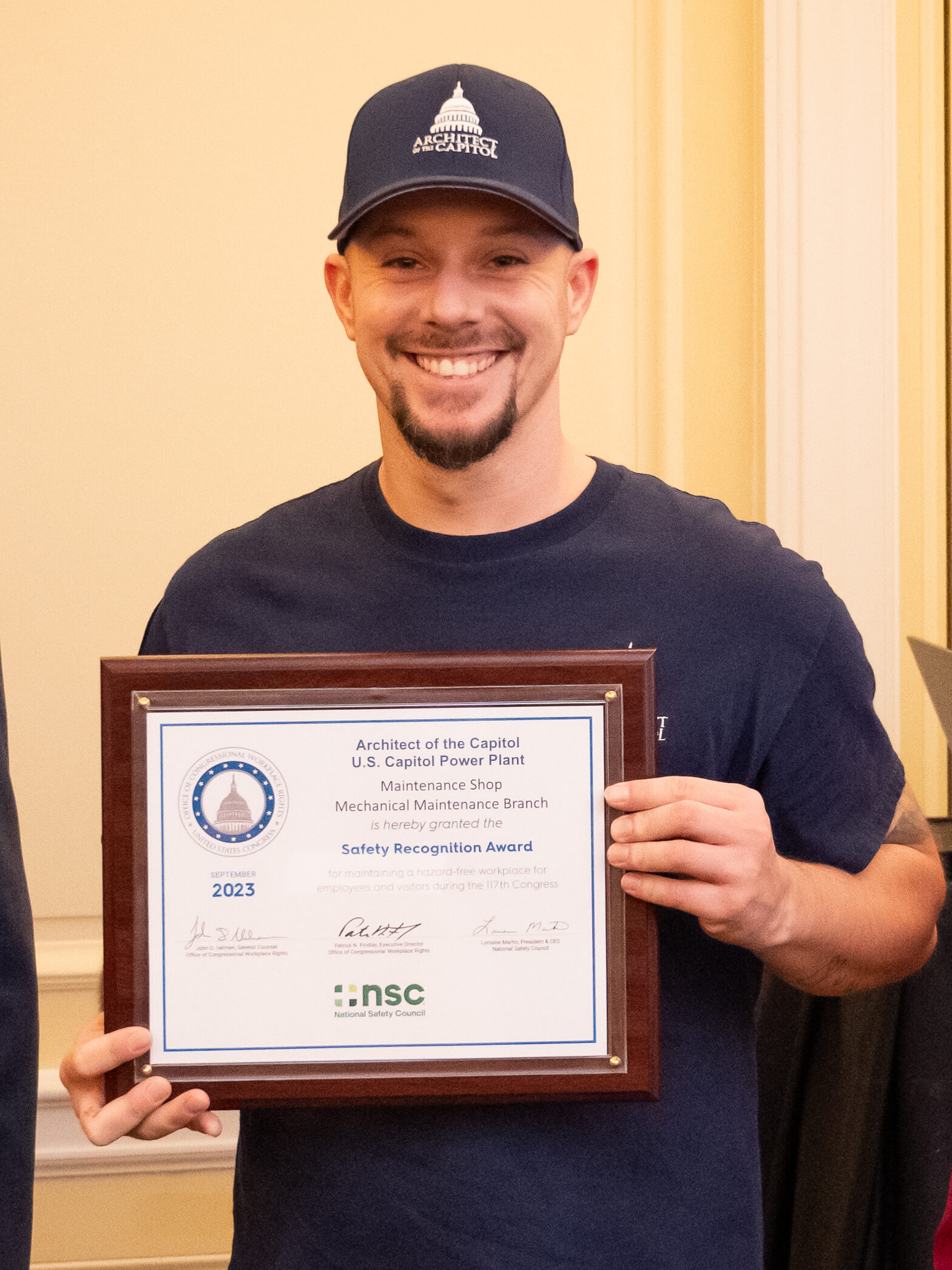 Man wearing a baseball cap with the Architect of the Capitol logo smiles holding a framed certificate