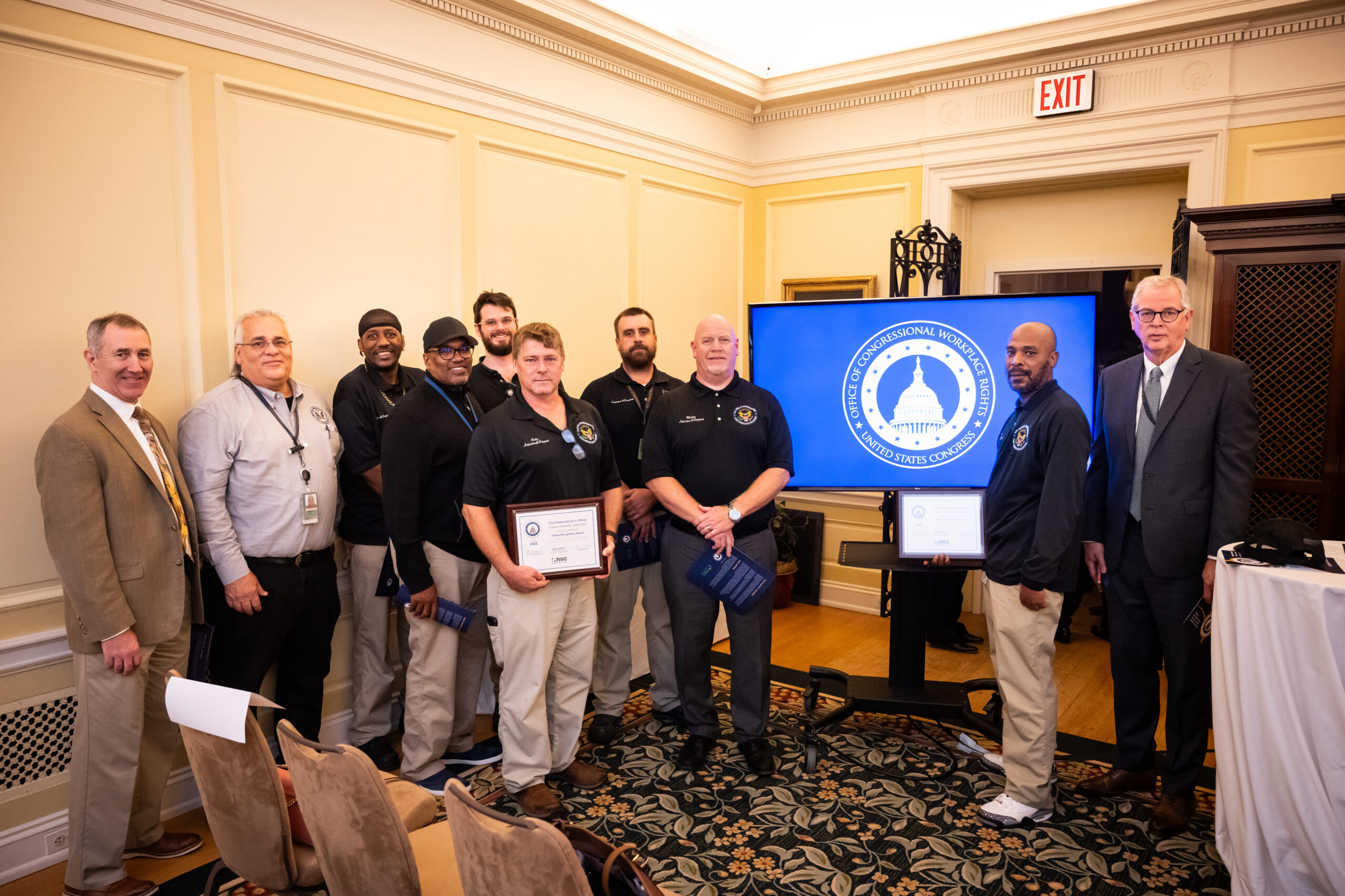 A group of ten men pose next to a screen displaying the Office of Congressional Workplace Rights logo