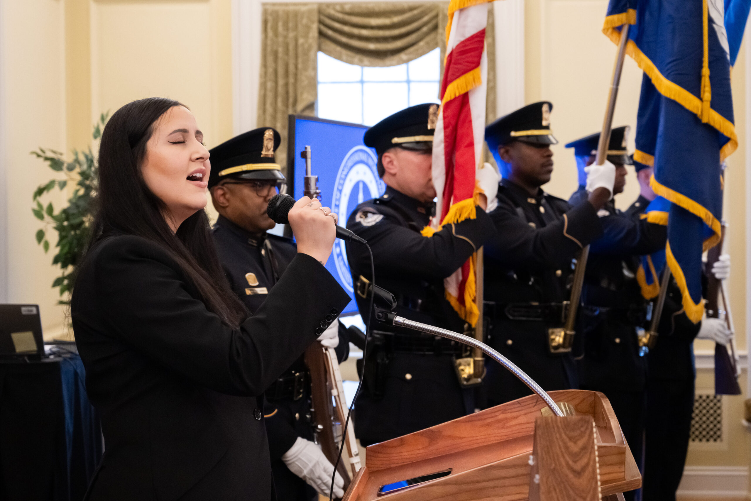 A woman sings into a microphone at a podium on the left with a police color guard standing behind her to the right