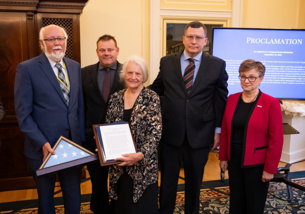 Three men and two women pose for a photo, the man on the far left holds a framed flag and the woman beside him holds a printed proclamation