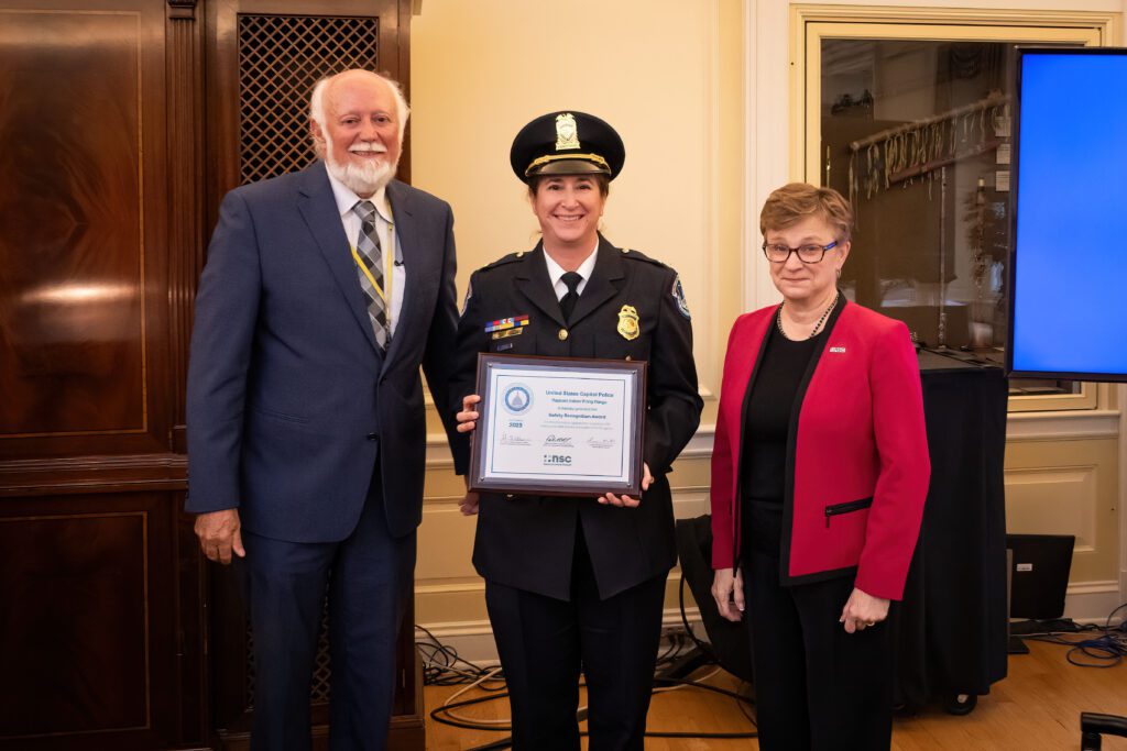 A female police officer in dress uniform holds a framed certificate with a man standing on the left and a woman on the right