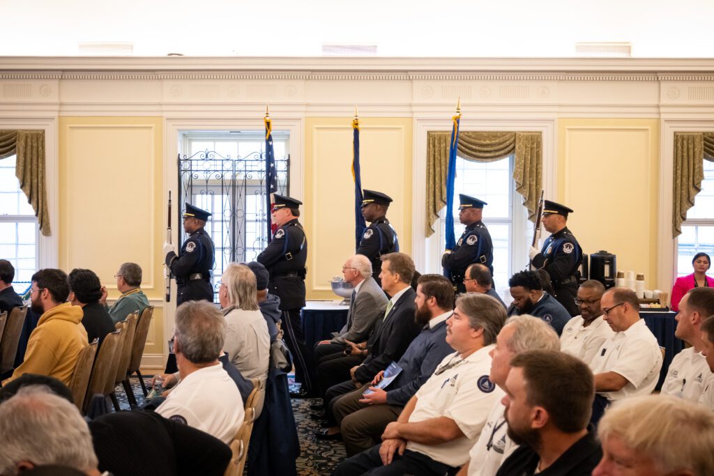 A police color guard marches along a wall with four windows in a room full of seated individuals